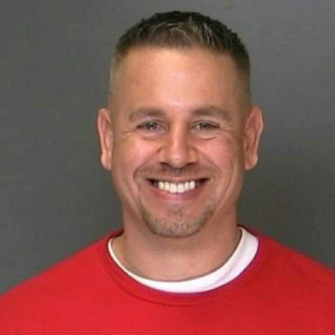 Stephen Ruth smiling broadly, wearing a red T-shirt over a white T-shirt, against a plain gray background